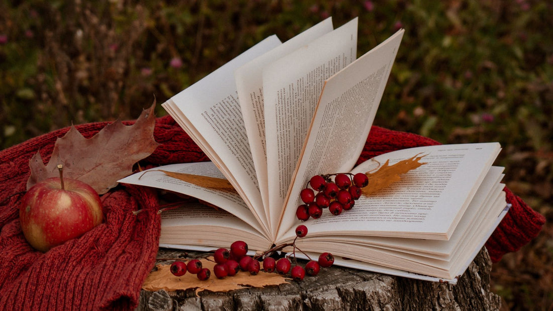 On a stump, the pages of an open book flutter in a breeze, adorned by fall berries and fallen leaves. Beside the book, a red knit material, and an apple.