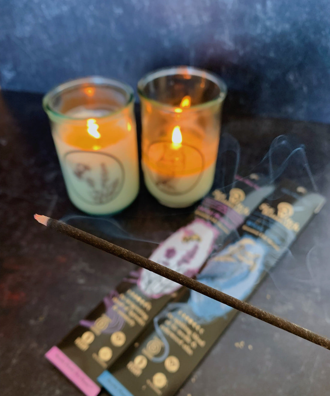 Incense stick burning in foreground, dark background with two lit candles and two packets of incense.
