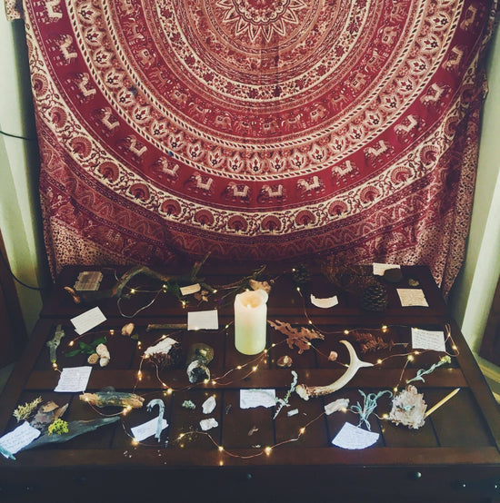 wooden table with scattered candles, fairy lights, antlers, flowers, papers, and leaves, red mandala tapestry in background