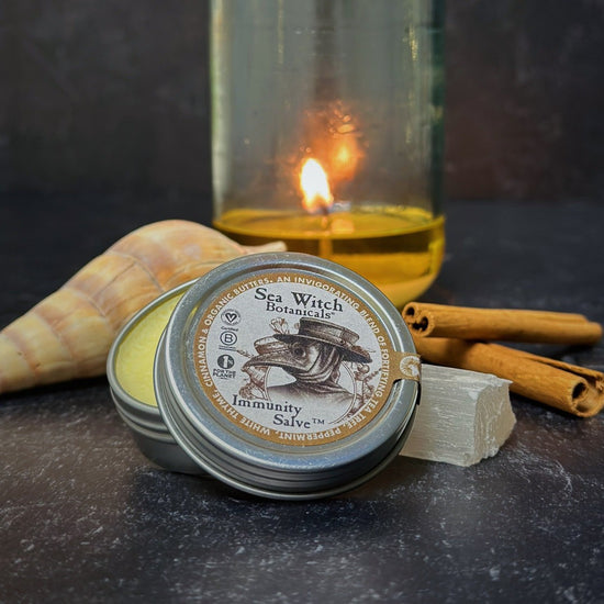 Immunity Salve from Sea Witch Botanicals. Inspired by plague doctors of the black plague. Pictured on a dark background with a shell, crystal, cinnamon sticks, and burning candle.