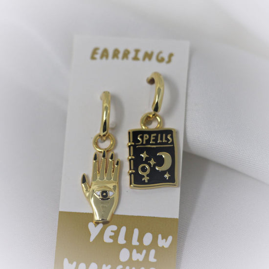 Yellow Owl Workshop Drop Charm Earrings - Mystic Spells. One is a golden hamsa hand and the other is a black book of spells. 