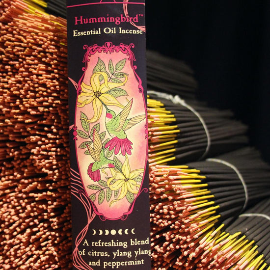A 20 pack of Hummingbird essential oil incense is posed against a stack of bundled Hummingbird incense sticks. (Close-up.)