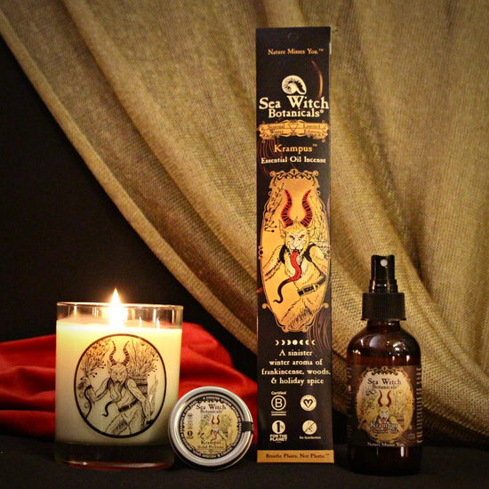 the 2022 limited edition krampus home and body scent collection, with candle, solid perfume, incense, and scented veil on a black set with red and gold fabrics in the background