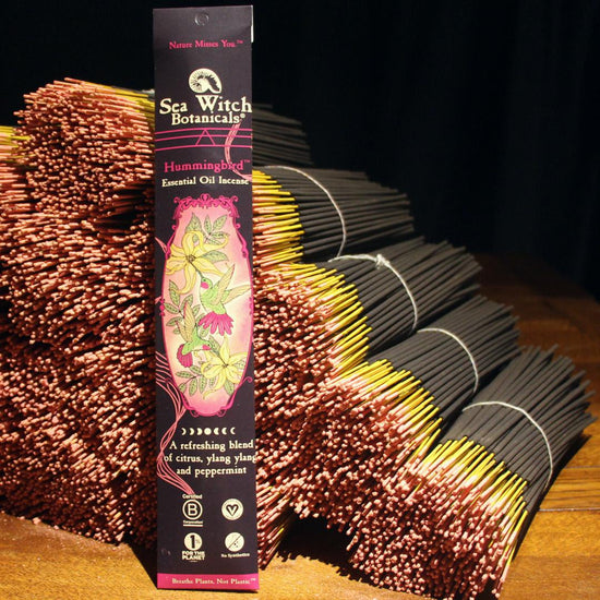 A 20 pack of Hummingbird essential oil incense is posed against a stack of bundled Hummingbird incense sticks.