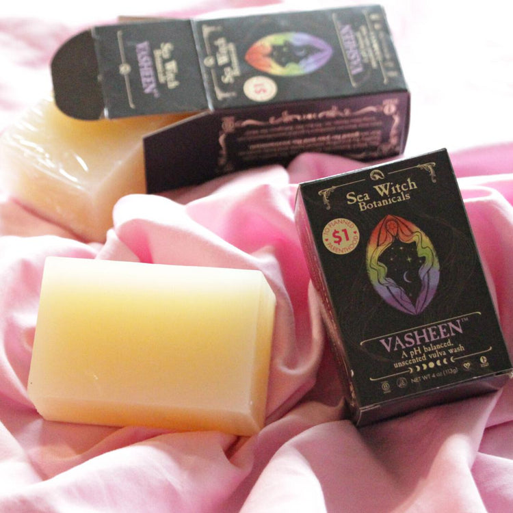 Vasheen vulva wash soap on pink fabric. In the background, one box is open with the bar halfway out. In the foreground, a naked bar sits beside a closed box displaying the Vasheen name and logo.