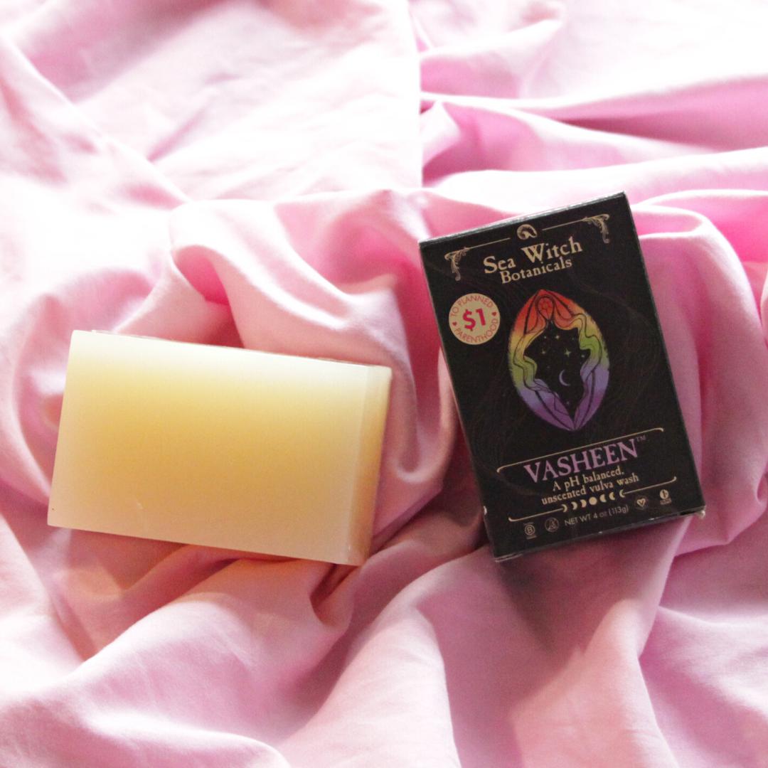 Vasheen vulva wash soap on pink fabric. A naked bar sits beside a closed box displaying the Vasheen name and logo.