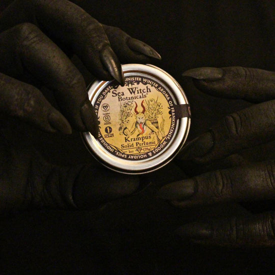 soot-black hands with claw-like nails holding a tin of 2022 limited edition all natural Krampus solid perfume