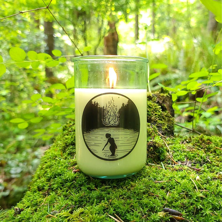 A Sea Witch Botanicals Limited Edition Litha candle sits atop a mossy log with green forest in the background.