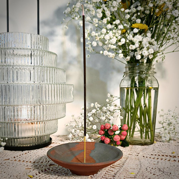 A stick of Ostara incense burns on a red clay incense holder on a lace covered table with flowers